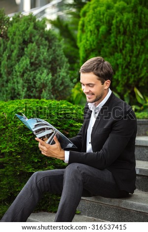 Fashionable man is sitting on stairs and reading a journal of luxury life. He is wearing a dress code like a rich man and smiling. He enjoys his life