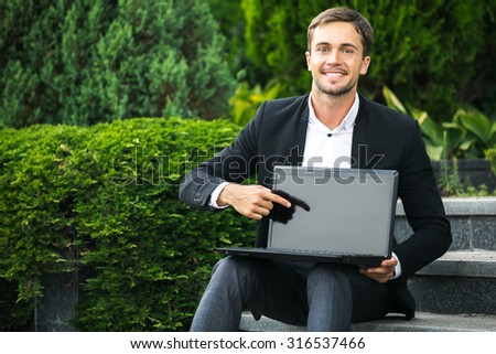 Young smiling man is sitting on the stairs in the park with green plants and pointing his finger to the laptop. He is showing something to us on his laptop. He can work everywhere he wants