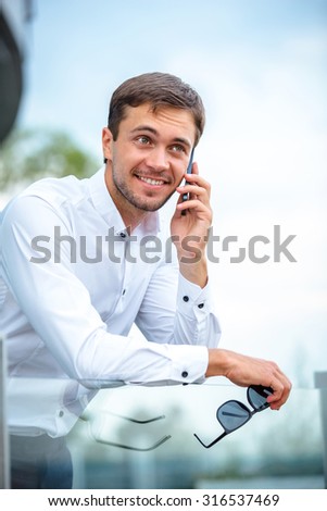 Fashion man in white shirt is standing with telephone. He is talking with someone. He is holding his glasses in hand. He is smiling