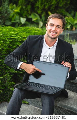 Young smiling man is sitting on the stairs. He is pointing his finger to the laptop. He is happy a lot