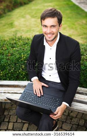 Young man is sitting on the bench with his computer. He was working on it. He is looking for us and smiling. He wears a white shirt and smart black suit.