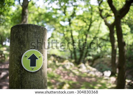 A black arrow on a light green background indicates progress on a footpath through a forest. Taken in the Peak District, Derbyshire, UK.