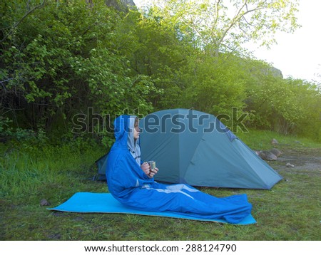 The man is resting in a sleeping bag on the background of tents and green forests.