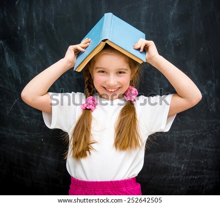 cute little girl with a blue book on her head smiling on the background of a school blackboard