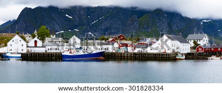 Fishing village on Lofoten islands. Steep hills in background partially covered in clouds. A large blue fishing boat with some seagulls around, wooden pier and smaller fishing boats in front.
