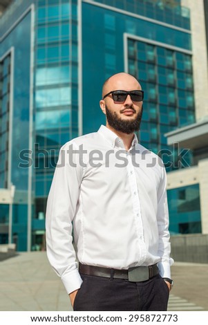 Businessman with beard in sunglasses in white shirt standing near office building