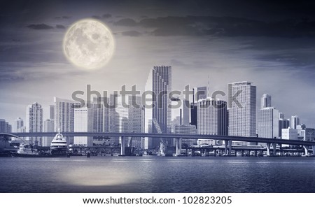 Dark color processed panorama of a full moon rising in the night sky over the city of Miami, with downtown buildings and Biscayne bay bridge.