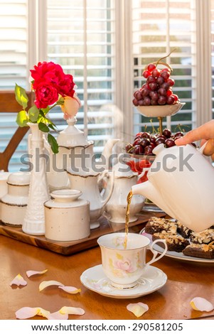 Afternoon tea table in rosy warm late afternoon sun. Black tea, fruits, brownie, cherries. Rose in cream white Depression Glass slim vase.