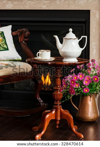 Afternoon tea at the fireplace. Antique armchair, wine table, copper vase, fire burning in the fireplace, vintage English teapot and teacup. Early retirement.
