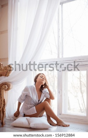 Portrait of charming girl in sweater sitting on windowsill and looking through window