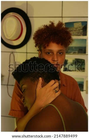 Portrait of a black LGBTQ+ couple hugging in a kitchen, with photos, hat and necklace in the background, on the wall. Imagine de stoc © 