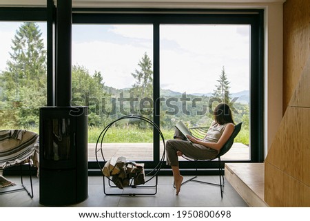 Beautiful stylish woman reading book on chair at fireplace with firewood on background of mountain hills. Young female in casual clothes relaxing in modern chalet with amazing view from window