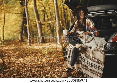 Travel and road trip with pet. Stylish happy woman traveller with cup and backpack sitting with cute dog in car trunk in sunny autumn woods. Young female traveling with swiss shepherd white dog