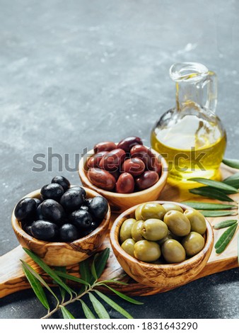 Set of green, black and red or pink olives and olive oil on gray background. Different types of olives in olive wooden bowls and olive oil over wooden cutting board. Copy space. Vertical