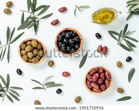 Set of green olives, black olives and red kalmata olives and extra virgin olive oil on white background. Top view of different types of olives in bowls and oil on leaves and branches background.