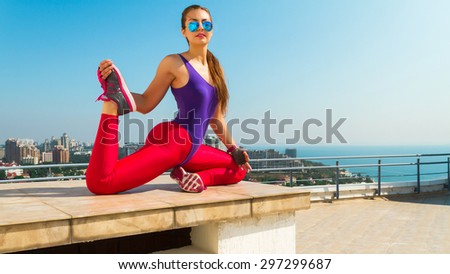 Sporty woman sitting in yoga position at sea view background.Healthy wellness lifestyle with multicultural young woman, wearing violet swimsuit,pink leggins, glasses.Making stretch after training.