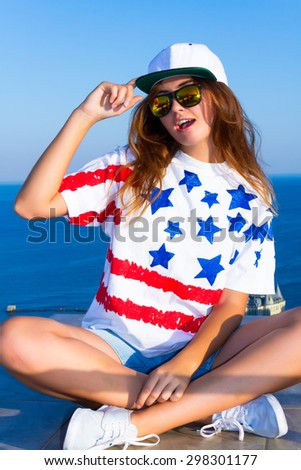 Summer cute portrait of young teen girl having fun and go crazy, wearing trendy top with american flag print,hat, hipster mirrored glasses. Sitting on the terrace with amazing sea view. Joy Sea Sunset