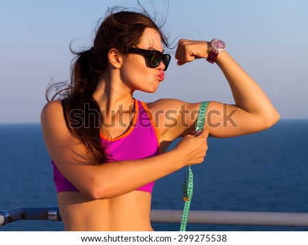 Fitness woman showing fresh energy flexing biceps muscles. Funny photo where girl what to kiss her biceps.Cheerfully smiling mixed race sporty woman demonstrating biceps with amazing sea background