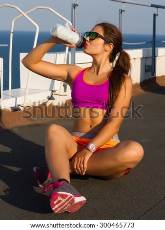 Fitness beautiful woman drinking water and sweating after exercising on summer hot day near the sea. woman athlete takes a break.Wearing bright purple sport bra,orange sport shorts,mirrored glasses