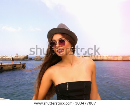 Summer fashion portrait of beautiful brunette woman enjoy sunny day near mediterranean sea,vacation,pin up style.Young stylish girl wearing black top corset,vintage grey hat and pink round sunglasses.