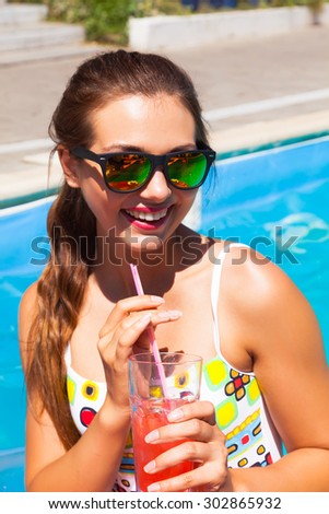 Outdoor summer lifestyle portrait of stylish sexy woman with perfect tanned fit body wearing trendy sunglasses drinking cocktail and enjoying pool party on luxury villa.