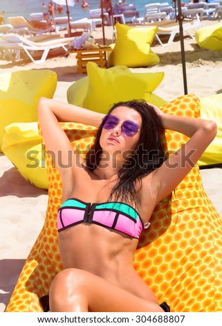 Summer fashion photo of beautiful tanned woman with brunette hair in sexy bikini relaxing at luxury beach resort on yellow beach chair. Summer close up of slim sexy woman having sunbath on vacation.