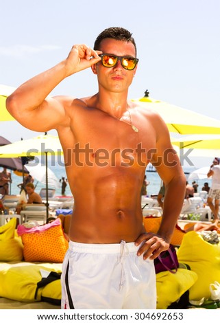 Summer close up fashion Portrait of a handsome young tanned muscular sexy man in white swim trunks and orange mirrored sunglasses with luxury beach resort background