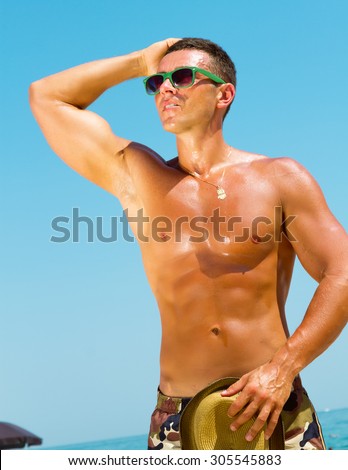 Sexy hot muscular Man on beach wearing hipster hat, sunglasses,military printed trousers.Young male model enjoying beach.