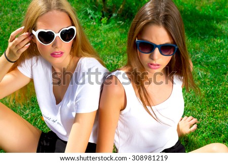 Two friends, stylish and young women in the park.Two hippie pretty young friends sitting on the grass. Girls take rest in city park. Both wearing vintage sunglasses,white t-shirt and black trousers