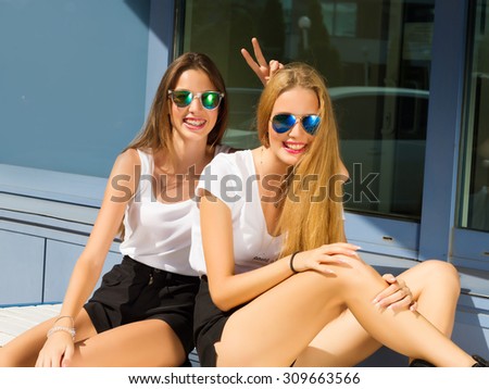 Two beautiful happy girls in sunglasses on the urban background. Young people. Outdoors.Pretty beautiful brunette and blonde girl friends in sunglasses resting and having fun in hot day.