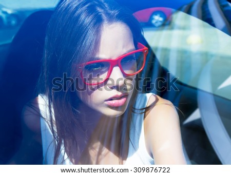 Young woman driving her car.Stylish student girl in car.Summer fashion close up portrait of elegant beautiful woman driving her car in urban city, wearing white shirt and red clear glasses.