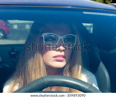 Young teenage woman driving her car.Stylish student girl in car.Summer fashion close up portrait of elegant beautiful woman driving her car in urban city, wearing black shirt and black clear glasses.