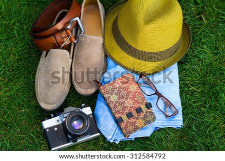 Spring or summer men\'s fashion accessories lying on grass, vacation concept for tourist.Men\'s casual outfits.Male accessories.Male hat,suede shoes, vintage camera,belt,notebook,clear glasses,and jeans