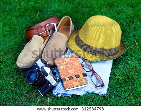 Spring or summer men\'s fashion accessories lying on grass, vacation concept for tourist.Men\'s casual outfits.Male accessories.Male hat,suede shoes, vintage camera,belt,notebook,clear glasses,and jeans