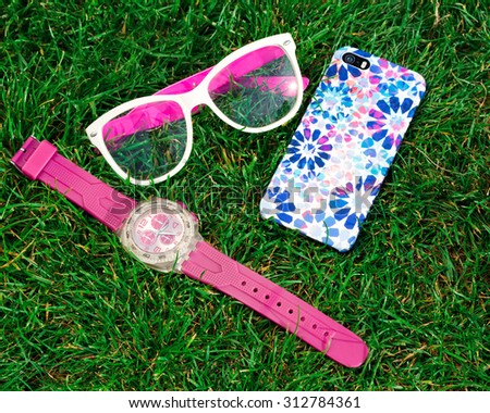 Women\'s fashion useful  accessories lying on grass. Pink watches,clear trendy glasses,and mobile phone with fashionable trendy cover on.