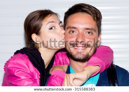 Night portrait of happy Couple at white urban wall background.Portrait of cheerful and lovely couple.So happy. Beautiful young lady giving kiss to cheek to his boyfriend with beard.