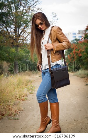 Autumn fashion image of young woman  walking in the park,wearing leather coat,white blouse,jeans and bag.Fashion urban young woman living city lifestyle walking in leather jacket,full length.