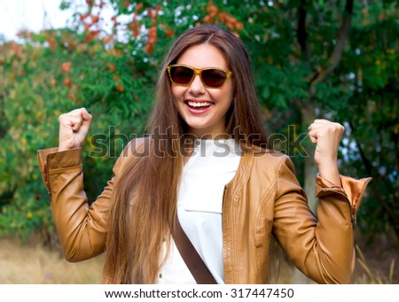 Autumn fashion close up portrait of young woman walking in the park and smiling,looking at camera, wearing leather coat,white blouse and sunglasses, raising his arms with a look of success
