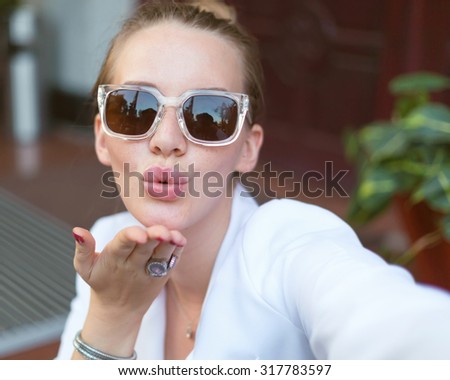 Positive selfie portrait of positive blonde girl sending kiss and  wearing white jacket and sunglasses.Close up portrait of sexy hipster girl sending kiss.trendy summer elegant outfit.