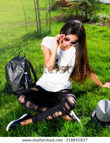 Trendy Hipster Girl Relaxing on the Grass.Modern Youth Lifestyle Concept.Happy beautiful teenage girl with long dark hair  sitting on grass in park on sunny summer day with leather bag and hat.