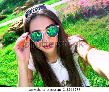 Young brunette woman in summer swag outfit walking in green park enjoying the sun and making selfie portrait. Playful and beautiful mixed race girl on warm sunny day.Student on holidays.
