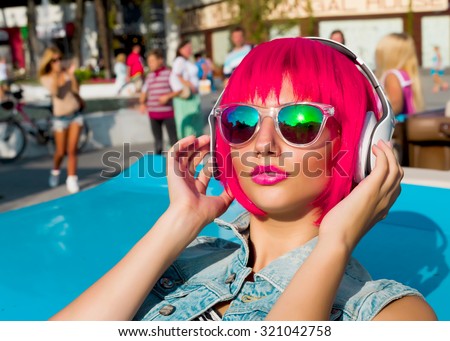 Close up fashion portrait of cute young girl with pink hair listening and enjoy music on headphones.Summer portrait of swag stylish sexy woman wearing hair wig, mirrored sunglasses, ready for party.