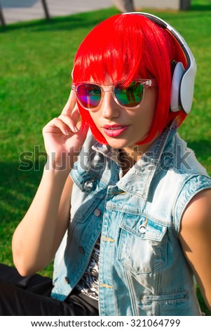 Close up fashion portrait of cute young girl with red hair listening and enjoy music on headphones.Fashion photo of sexy beautiful girl with red wig hair in jeans vest. Outdoors lifestyle portrait.