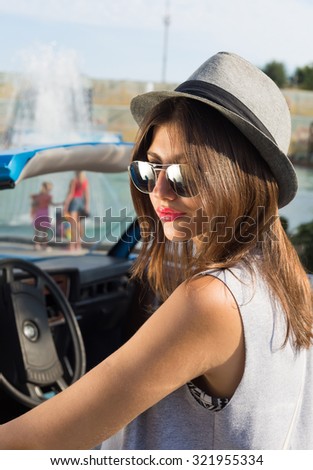 Young woman standing by car,beautiful bright sunny summer day. Summer vacation car road trip freedom concept.Teenage hipster girl wearing hat,sunglasses and fashionable dress.Smiling,positive.