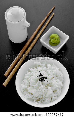 A dish of steamed rice with chopstick, soy sauce jug and wasabi paste on a black background