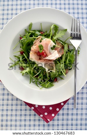 A rocket salad with parma ham slices and pomegranite seed in a white bowl on a blue checked table cloth with a red and white polkadot napkin