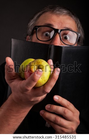 Old man with beard and big nerd glasses with apple in head showing apples in hand hides   behind big sleek leather book