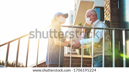 Female courier in medical mask and gloves bringing packet with fresh food to retired old man. Young Caucasian woman handing bag with grocery to male pensioner outdoor at house. Delivery service 商業照片 © 