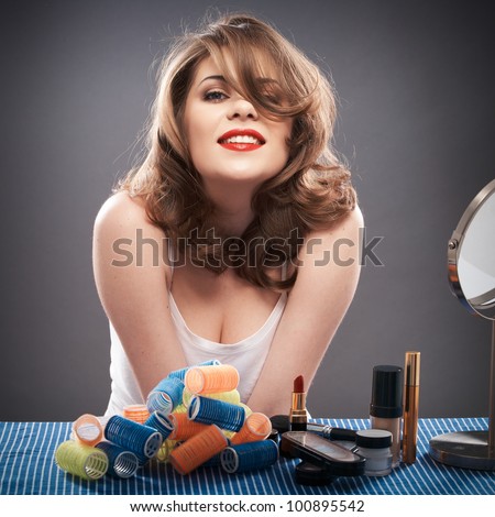 Portrait of a young woman with long hair on gray background isolated.  Happy girl seating at table with make up accessories and mirror. Smiling model with curler hair dress