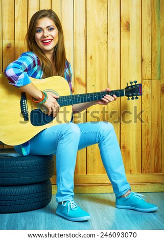 Girl hold guitar, sitting on a car  wheels against wooden background.